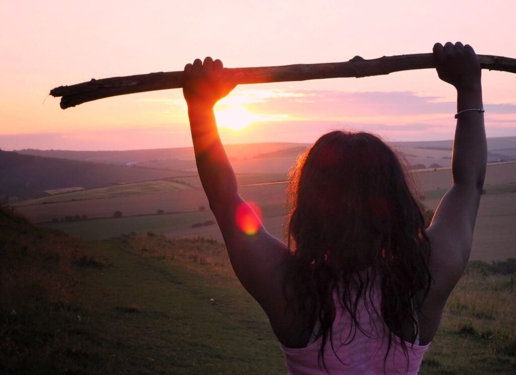 Girl holds a stick high in the air above her head with both hands, overlooking a valley