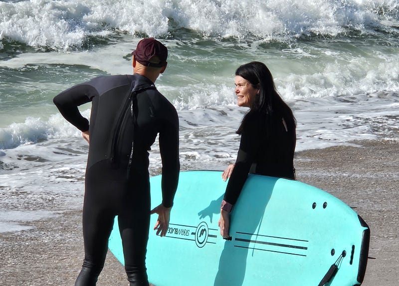 An instructor and woman holding a surfboard, stand on the beach. Surf in the background.