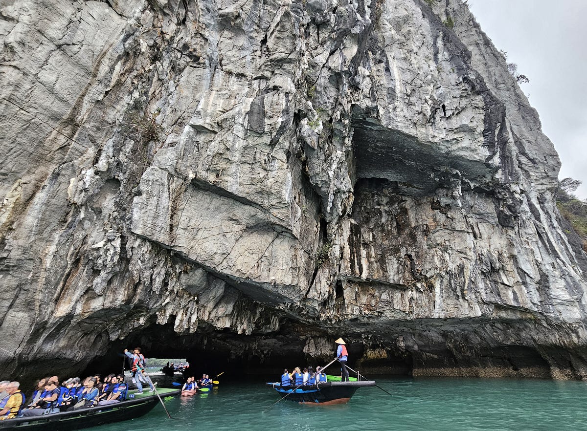 A large jagged rock formation, with small boats and kayaks floating under a tunnel carved by the water, under a karst.