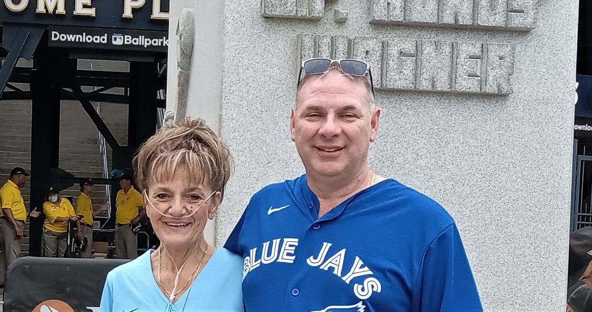 A woman with an oxygen canal stands with a man in front of the Pittsburgh Pirates ballpark. Both wearing BlueJay gear.