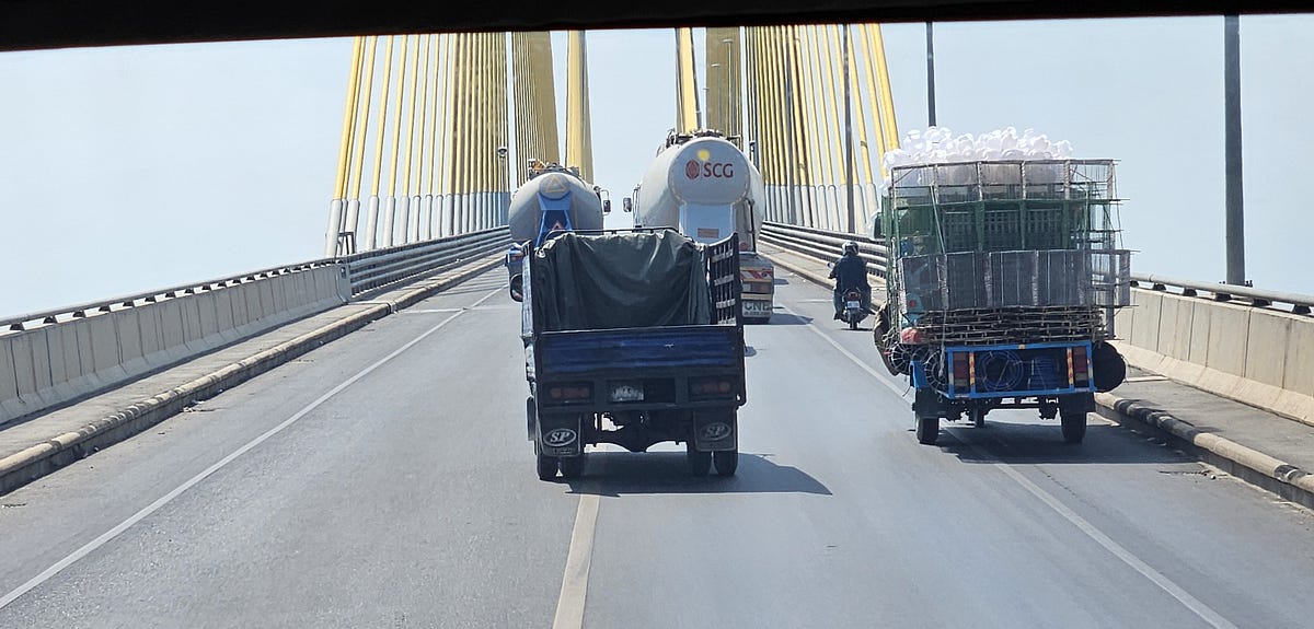 Large and small trucks go passed a heavily loaded motorcycle and trailer while climbing the grade of a tall, two-lane bridge.