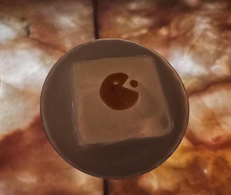 A drink sits on a marble bar. Pac-man has been cut from an orange for a nifty garnish, sitting atop a solid cube of ice.