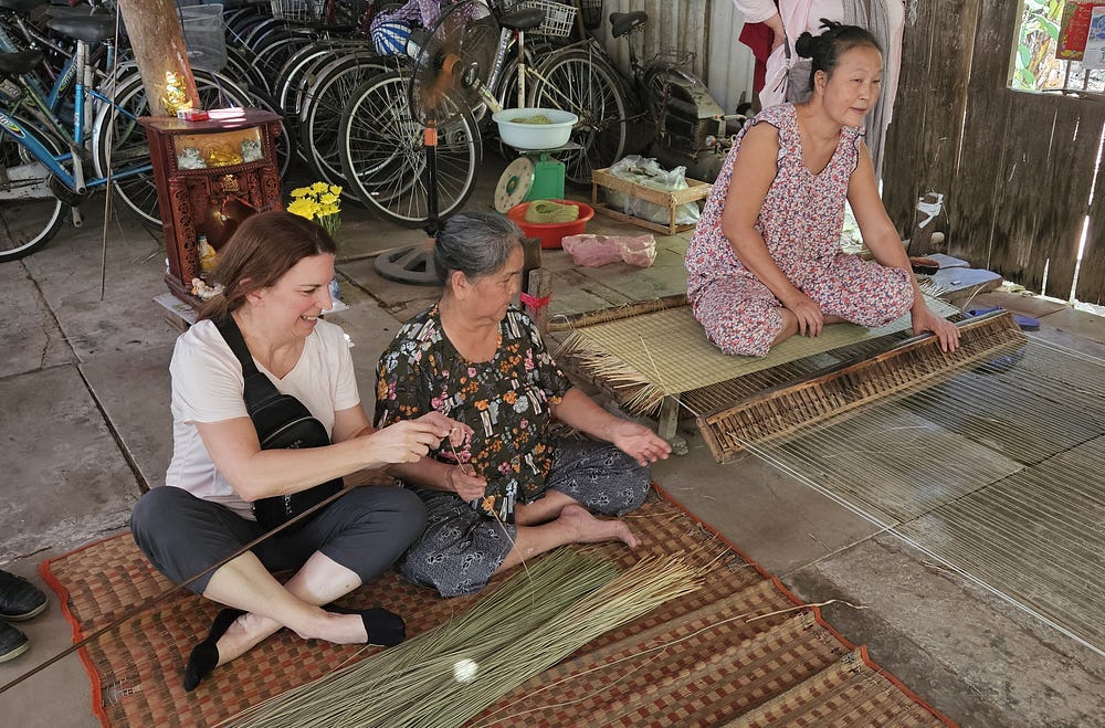 2 elderly Vietnamese women show a traveller how to weave a sleeping mat.