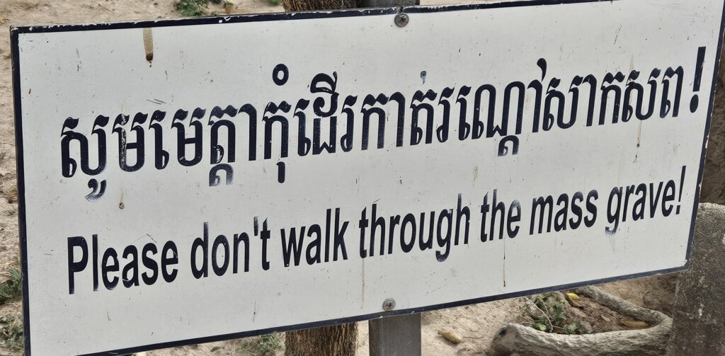 White sign with black lettering in 2 languages. 'Please don't walk through the mass grave!